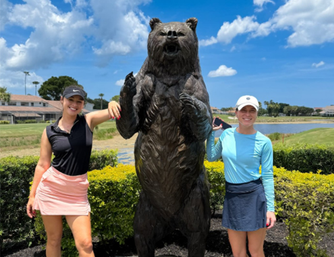 two golfers pose with large bear statue on golf course
